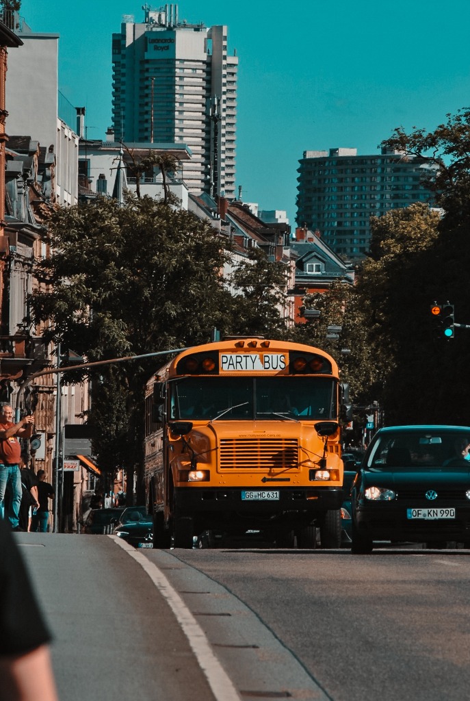Yellow bus driving down city street framed by skyscrapers by Tony